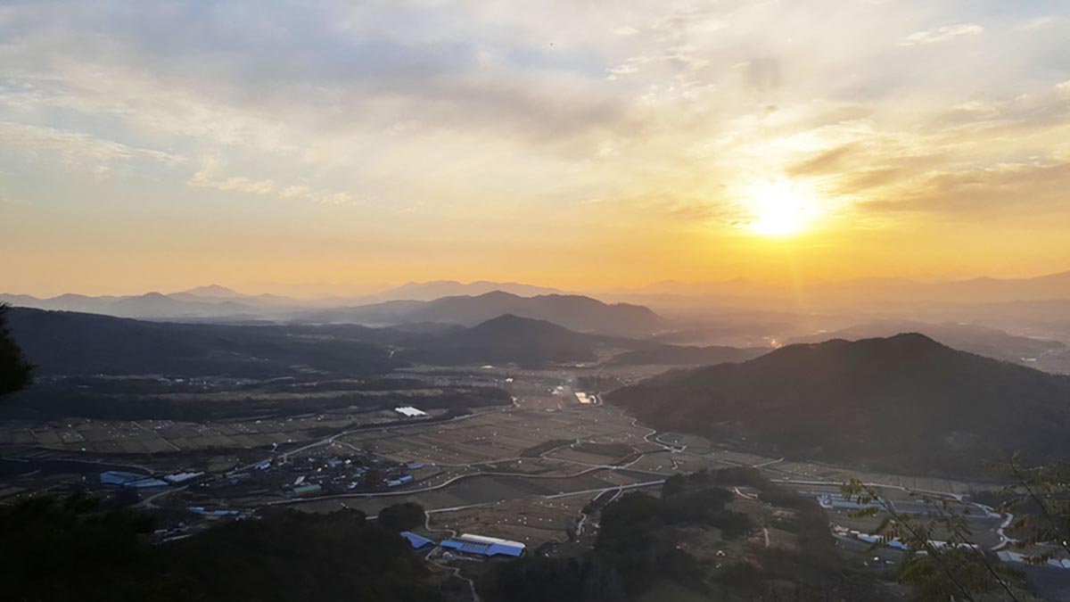 장수 안양마을을 높은 곳에서 찍은 마을 전경 사진입니다. 작은 마을이 겹겹이 펼쳐진 낮은 산들을 둘러업고 있는 모습입니다. 마을 중앙에는 여러 채의 민가들이 모여 있고, 그 주변으로 초록빛 논과 밭이 둘러싸고 있습니다. 하늘에는 주황빛 노을이 지는 모습입니다.