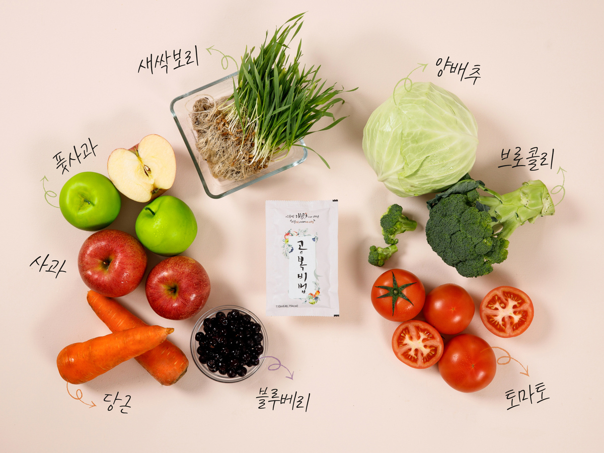 흰색 식탁 위, 중앙에는 흰색 직사각 비닐포장재에 포장된 공복비법 한 포가 올려져 있고, 그 주변으로 원재료들이 둥글게 놓여있습니다. 좌측부터 나열해보겠습니다. 둥근 유리그릇 가득 담긴 작고 둥근 검은색 아로니아 알갱이, 길쭉하고 큰 주황색 당근 두 개, 빨갛고 윤기나는 사과 두 알, 초록빛 풋사과 두 알, 연갈색 잔뿌리를 가진 초록빛 새싹보리 더미, 연두 잎사귀의 크고 둥근 양배추, 진한 초록색의 큰 브로콜리, 빨갛고 둥근 과실에 초록 꼭지가 달린 토마토 여러 알. 통째 올려진 토마토들 옆에는 반으로 가른 반쪽 토마토 두 개도 올려져 있습니다.