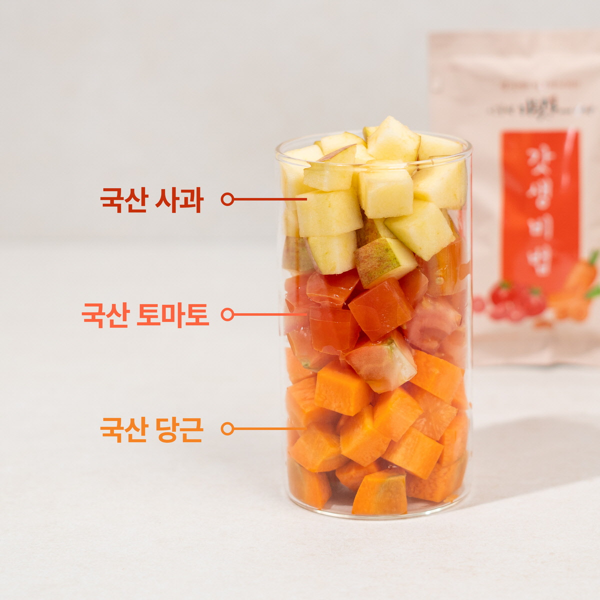 투명한 컵에 담긴 국산 사과, 국산 토마토, 국산 당근의 층층이 쌓인 모습. 갓생비법 과채주스의 신선한 원재료.