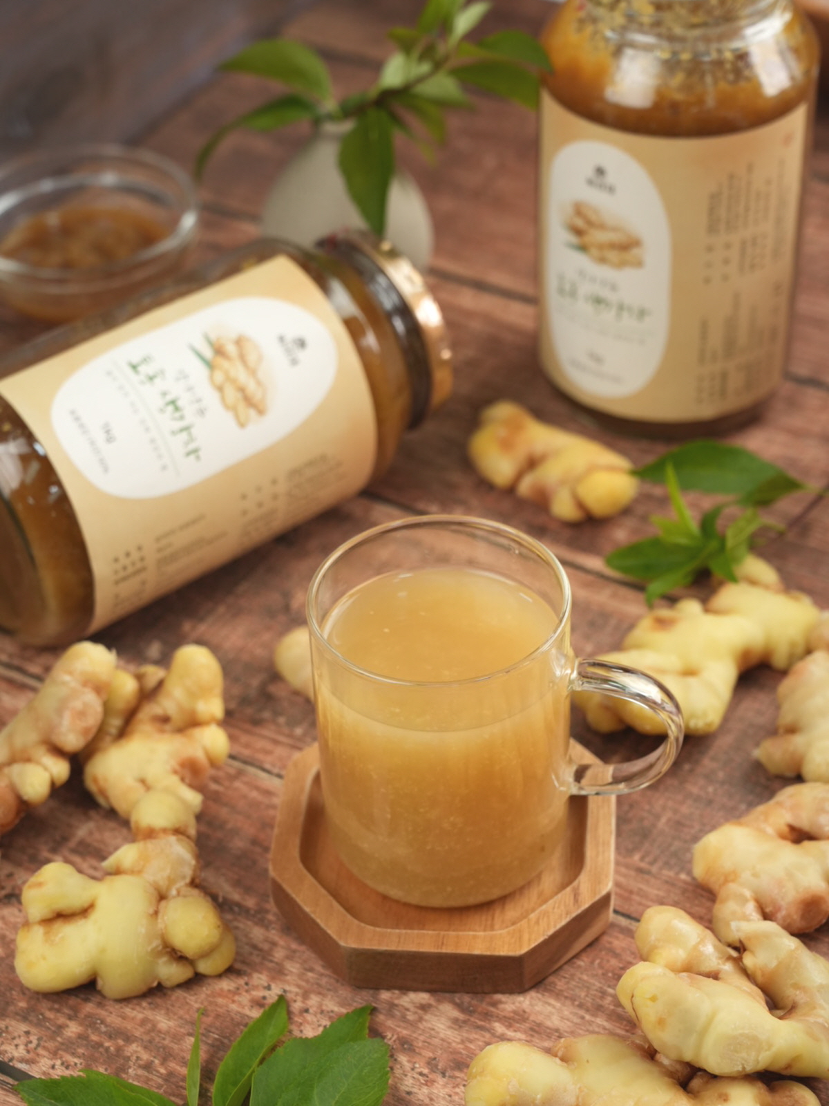 따뜻한 국산생강차 한 잔을 유리잔에 담아 손으로 들어올리는 모습. 생강청과 진저라떼에 활용 가능한 진한 색감의 음료가 나무 쟁반 위에 놓여 있고, 뿌리 생강과 허브가 함께 배치됨.